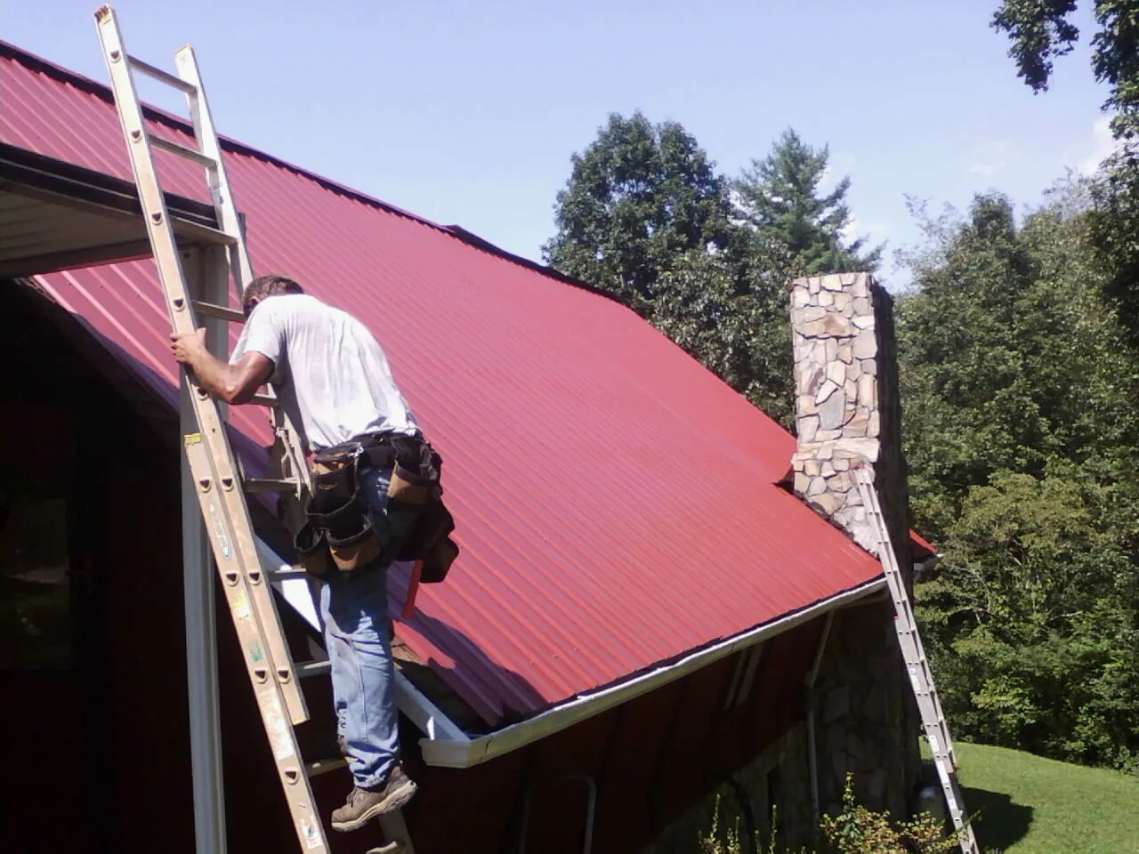 Roofer on a ladder beside a red metal roof and stone chimney — Soffit & Fascia Repair by Roofing Brothers in Kenoza Lake